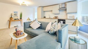 A modern living room with a grey sofa, a small white side table, and a kitchen area with white cabinets and a microwave. - Host Stay Apartment 1 Dane Court (Harrogate)