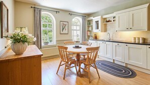 A kitchen with white cabinets, a wooden dining table, and chairs. There is a window with curtains, a vase with flowers, and a rug on the floor.