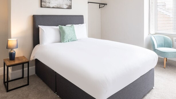 Een moderne slaapkamer met een groot bed, een nachtkastje met een lamp, een stoel en een raam met jaloezieën.