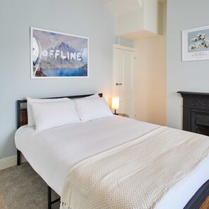 A neatly made bed with white linens and a textured throw blanket, a framed artwork above the bed, a white door, and a fireplace with a mantel. - Host Stay Wild Springs (Wetherby)