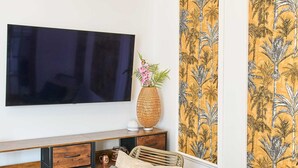 Un salon moderne avec un téléviseur à écran plat fixé au mur, un fauteuil à bascule en osier avec des coussins, une petite table ronde ornée d’un vase décoratif et un meuble en bois avec des tiroirs.
