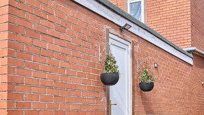 Un bâtiment en briques rouges avec une porte blanche, deux jardinières suspendues et un petit espace jardin avec des plantes.