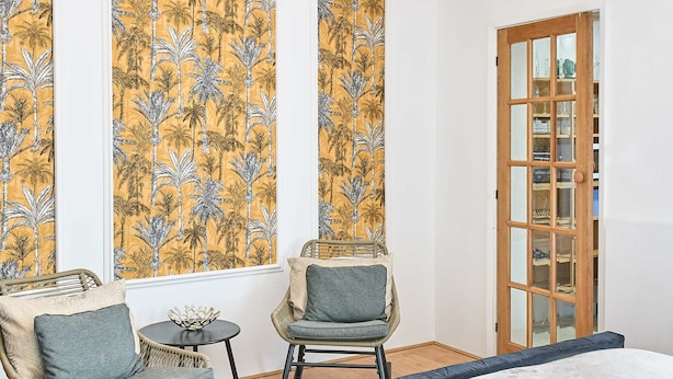 Une pièce comprenant un lit, deux fauteuils à bascule, une petite table et une fenêtre ornée d’un papier peint aux motifs tropicaux.
