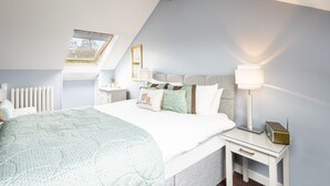 Ein Schlafzimmer mit geneigter Decke, einem Oberlicht, einem Bett mit Quiltdecke, Nachttischlampen, einem Nachttisch und einem Heizkörper.