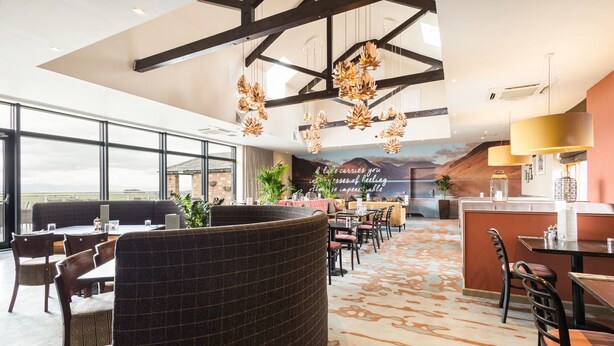 Ein modernes Restaurant mit hohen Decken, großen Fenstern und einer Wandmalerei einer Berglandschaft.
