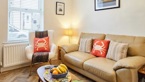 A living room with a beige sofa, a white armchair, a wooden coffee table with a bowl of fruit and a book, a window with blinds, a chandelier, and a framed picture on the wall. - Host Stay Sunstar (Whitby)