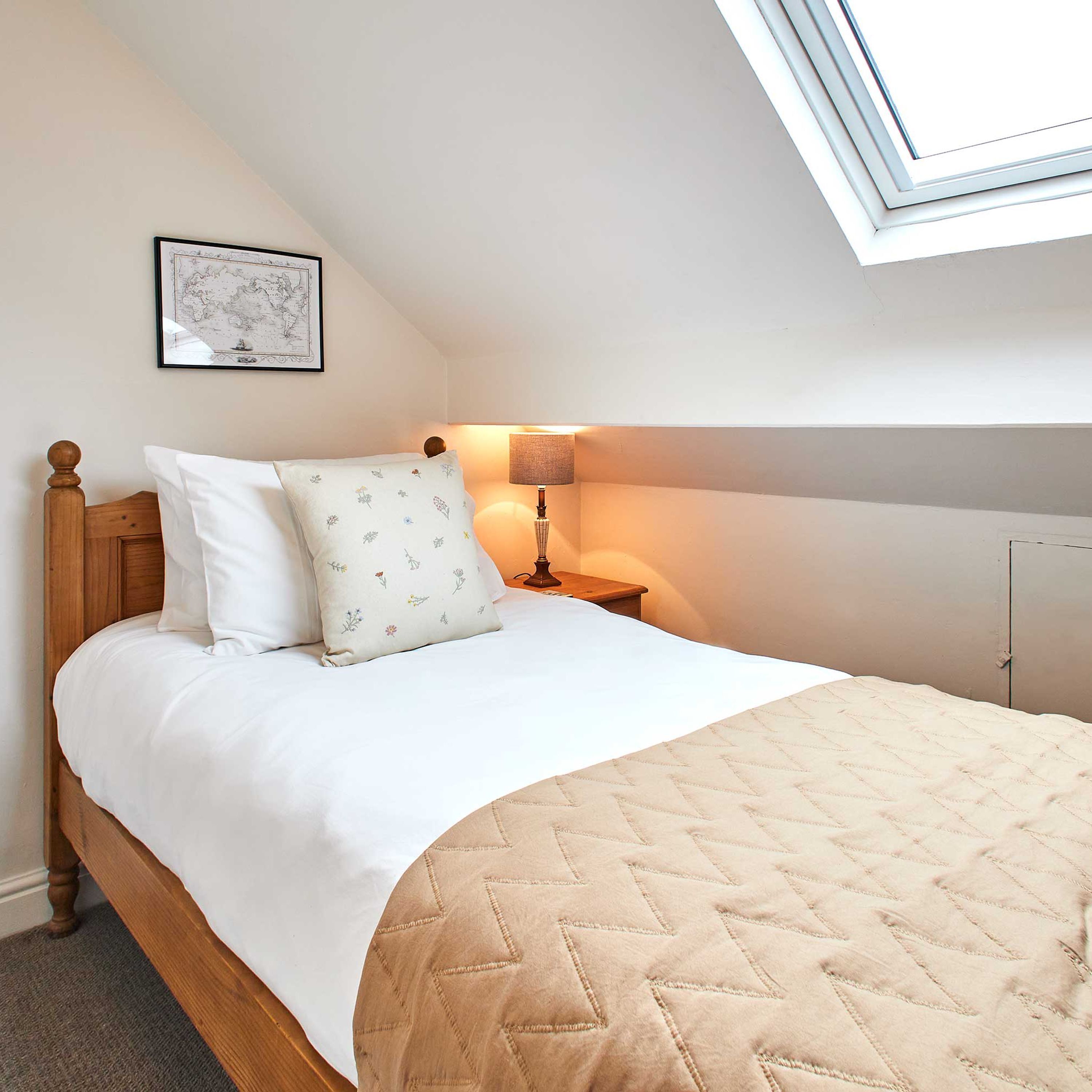 A bedroom with a wooden bed, a quilted beige bedspread, a white pillow with floral patterns, a bedside lamp, and a framed map on the wall.