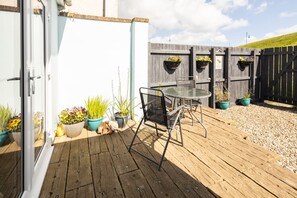 Um deck de madeira com mesa e cadeiras de vidro, cercado por vasos de plantas e um cercado de madeira.