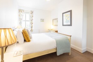 Quarto com cama, mesa de cabeceira, abajur, janela com cortinas, radiador e um quadro na parede.