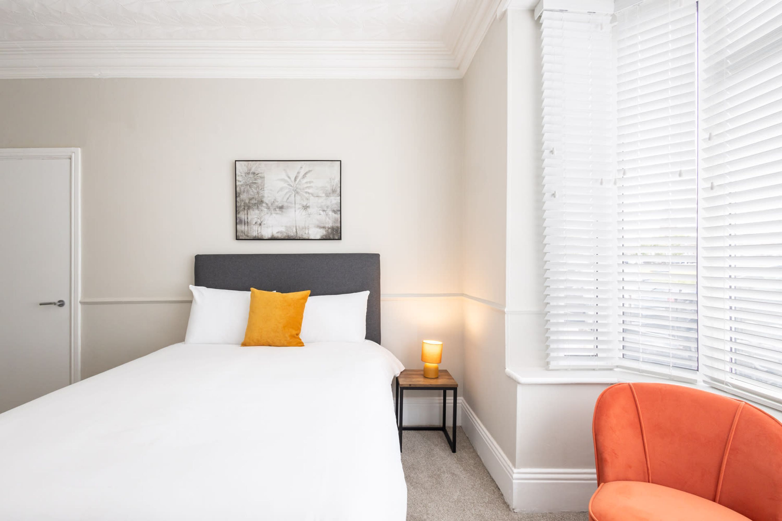 A bedroom with a bed, a bedside table with a lamp, a chair, and a window with blinds.