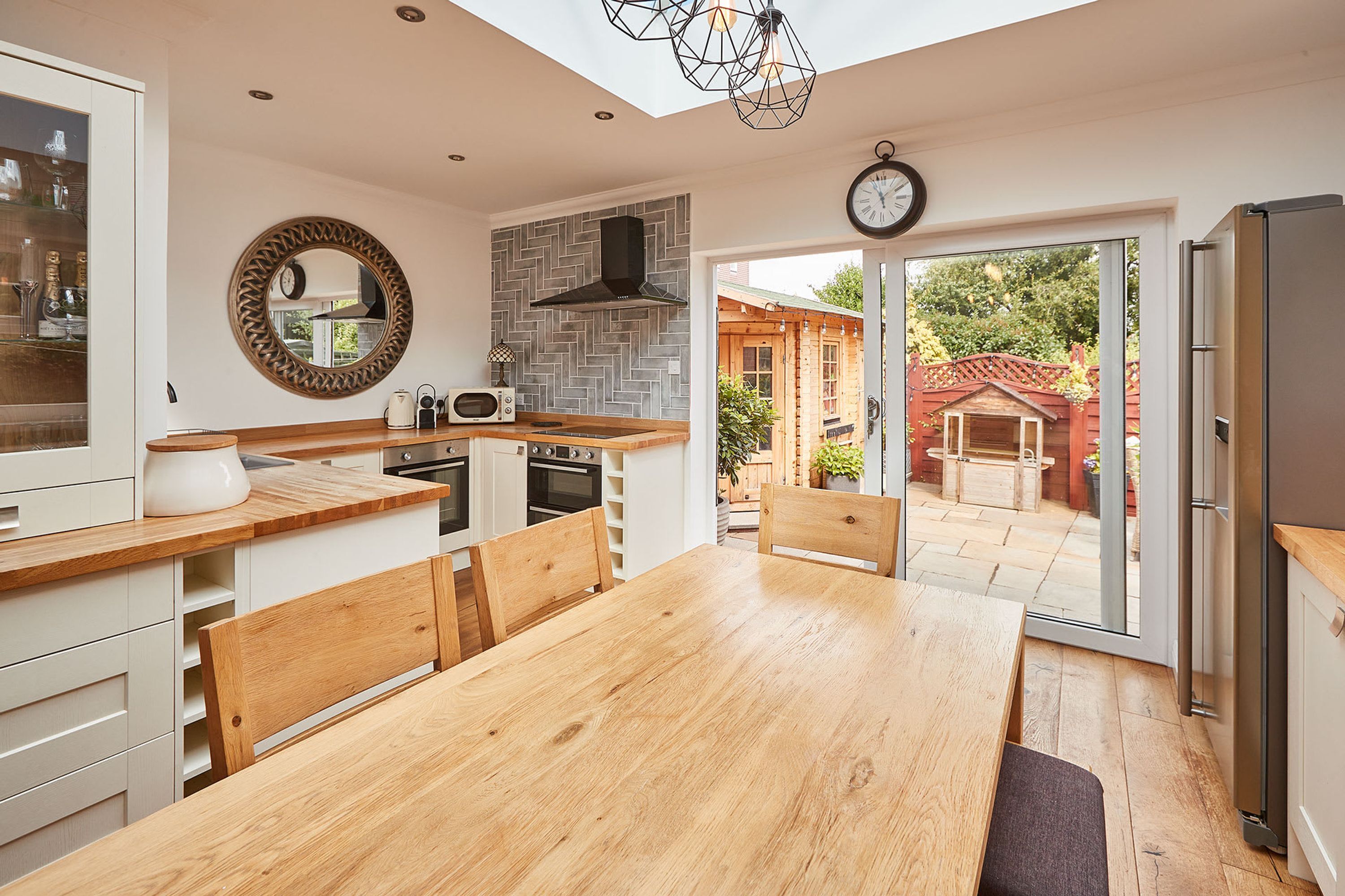 A modern kitchen with a wooden table and chairs, a built-in oven, and a large window overlooking a garden.