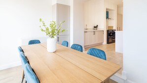 Ein moderner Essbereich mit einem Holztisch und blau gepolsterten Stühlen, einer weißen Vase mit Grünpflanzen und einer Küche im Hintergrund.