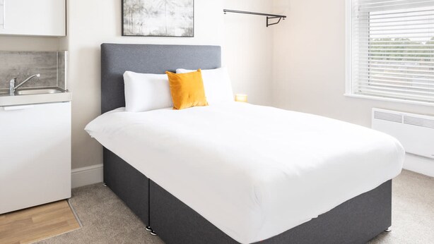 A modern bedroom with a large bed, a bedside table, a nightstand, a window with blinds, a wall-mounted shelf, and a kitchen area with a sink and countertop. - Host Stay Clifton Studio 4 (Darlington)