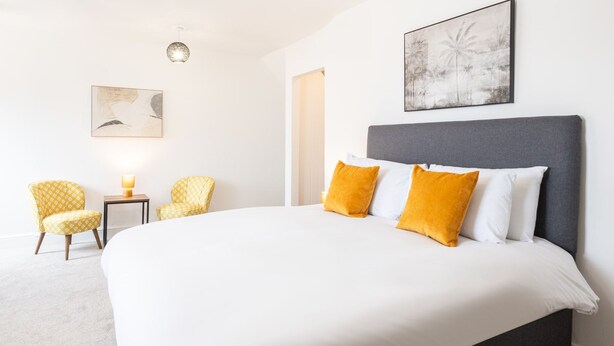 Una habitación de hotel moderna con una cama grande, dos sillones amarillos con estampado y una mesita de noche con lámpara, además de obras de arte en las paredes.