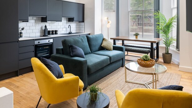 A modern kitchen with dark cabinets, a white tiled backsplash, and a built-in oven. There is a green sofa with cushions, a yellow armchair, and a round glass table with a plant. - Host Stay Princes Place Hideaway I (Liverpool)
