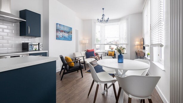 A modern kitchen with white cabinets and a built-in oven. There is a dining area with a round table and white chairs. - Host Stay Sanderling Chase (Scarborough)