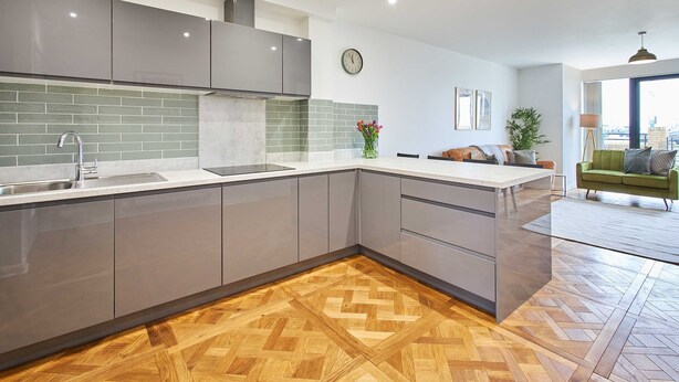 Cozinha moderna com armários cinza, piso de madeira em padrão herringbone, e vista para a sala de estar com sofá verde e uma planta.