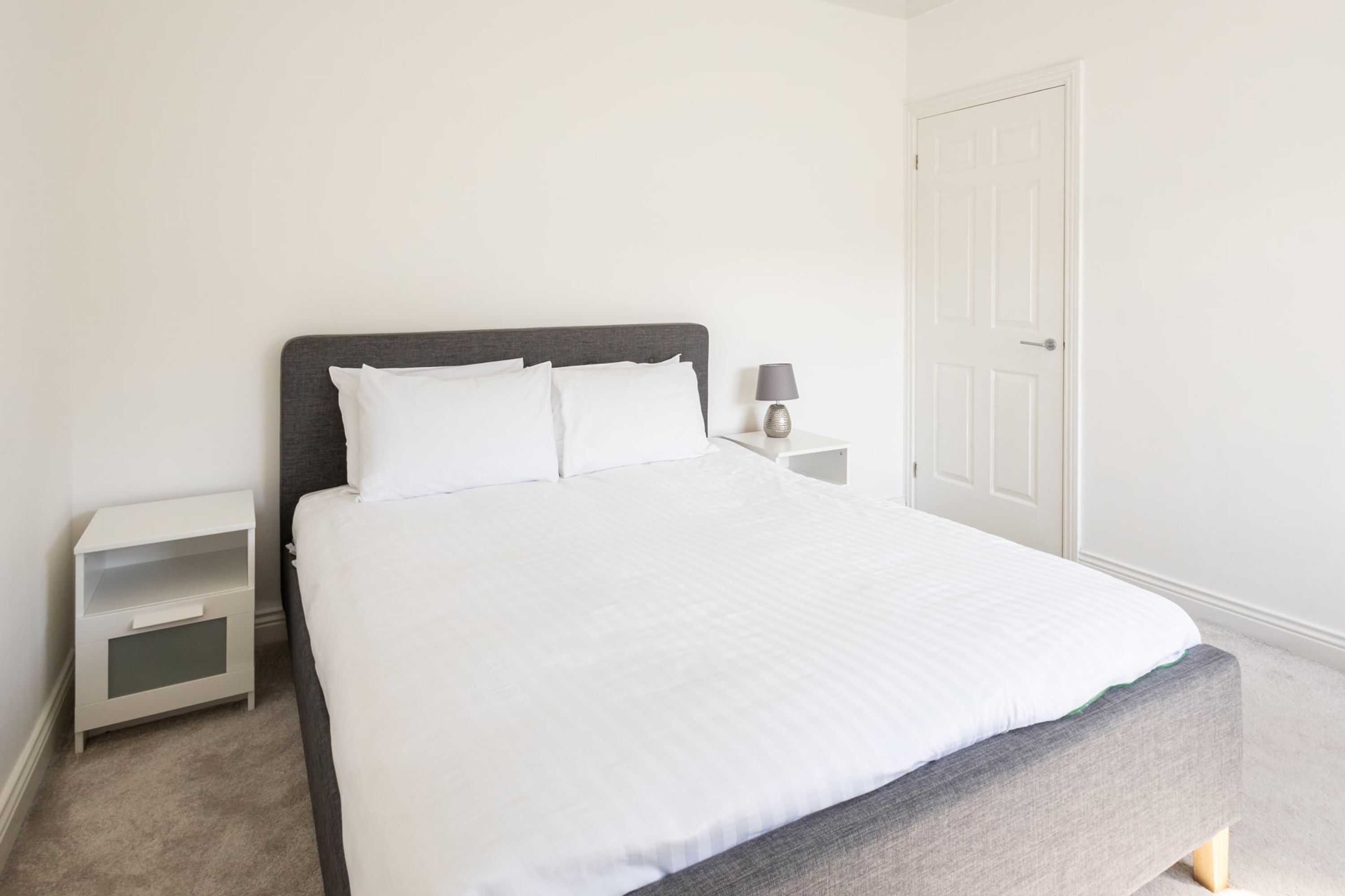 A neatly made bed with a grey headboard and white bedding, a bedside table with a lamp, and a white door in a minimalistic room.