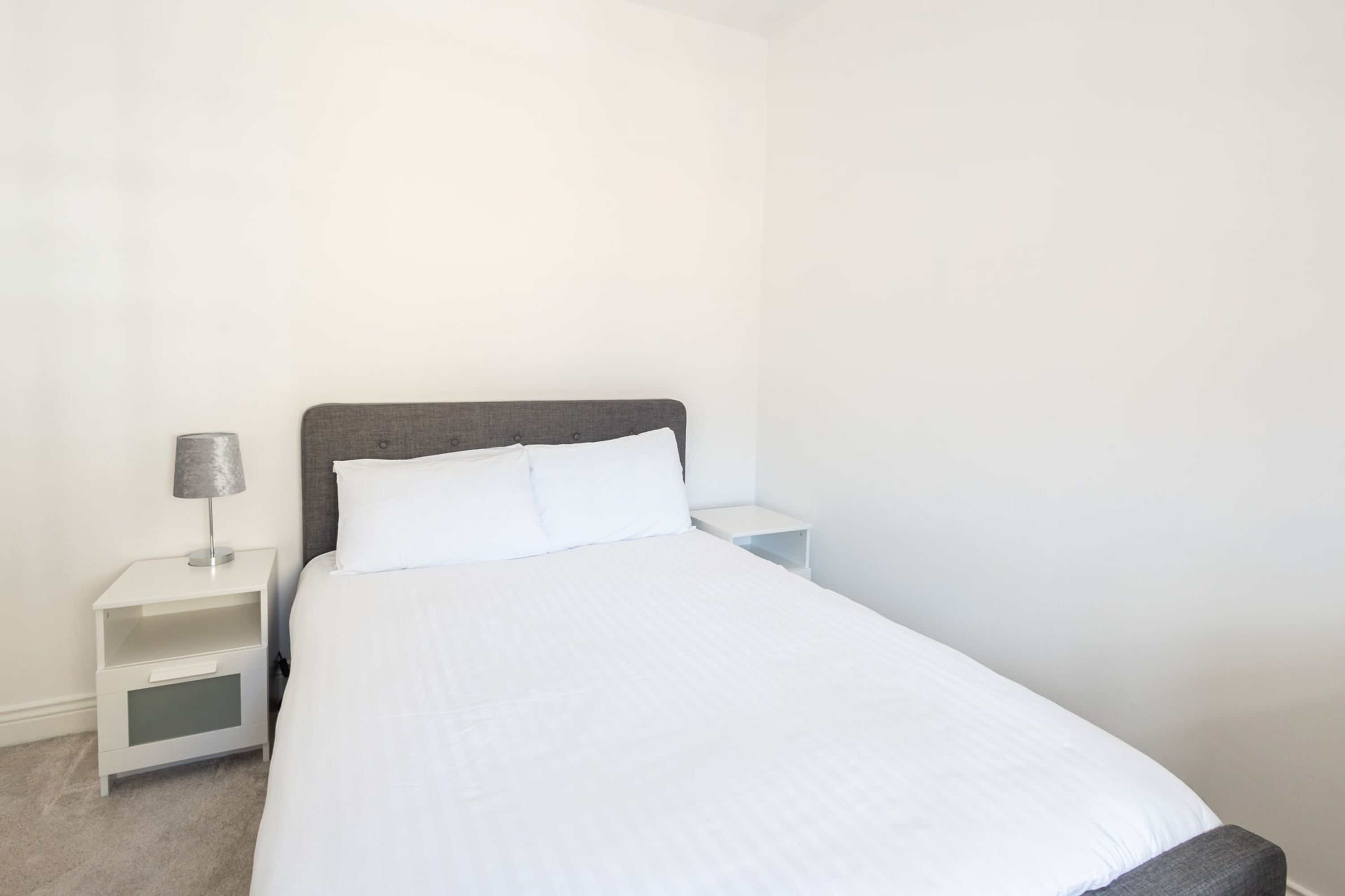 Una cama bien tendida con ropa de cama blanca y un cabecero gris en una habitación sencilla y limpia.