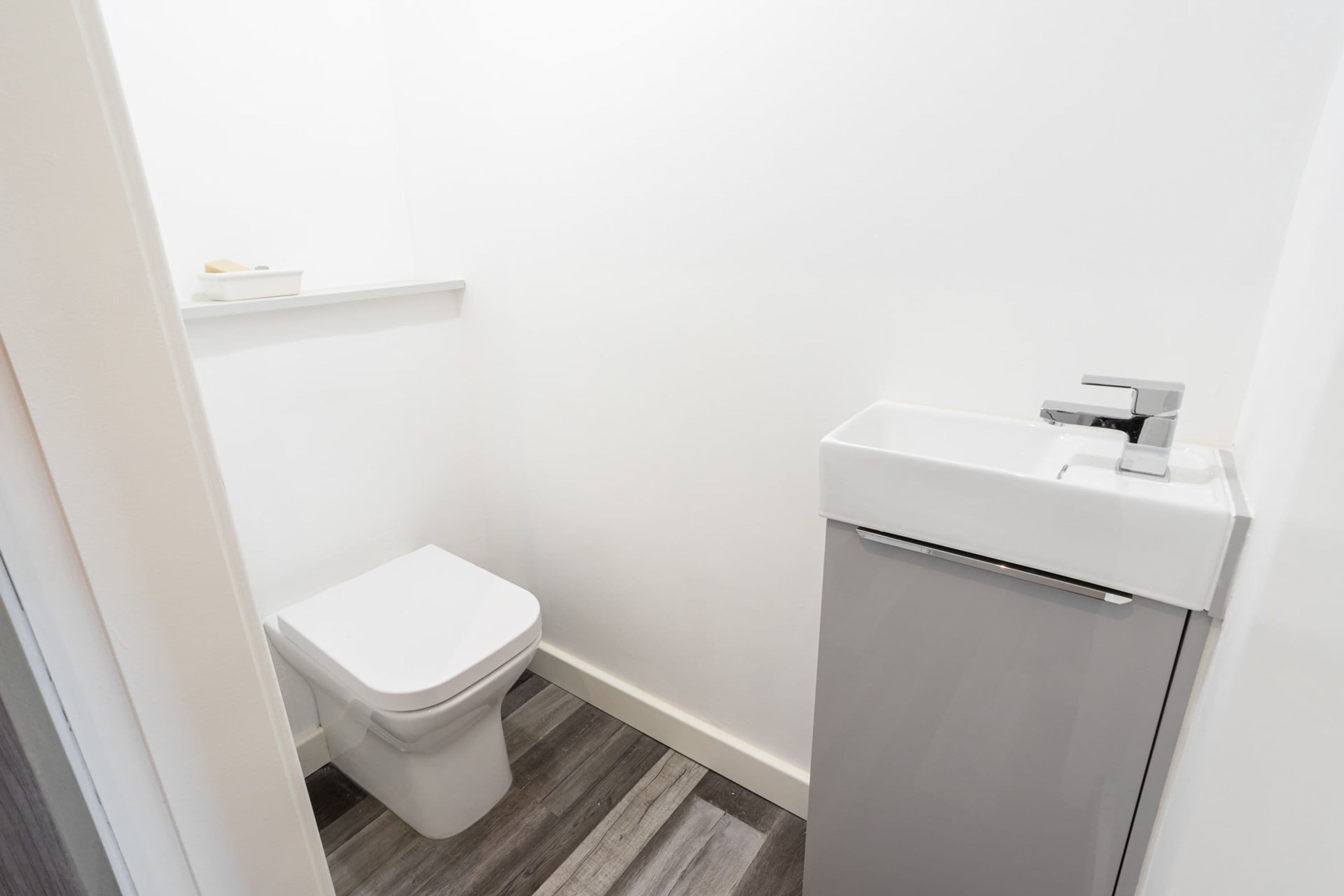 A small, clean bathroom with a toilet, a white sink, and a shelf above the toilet.