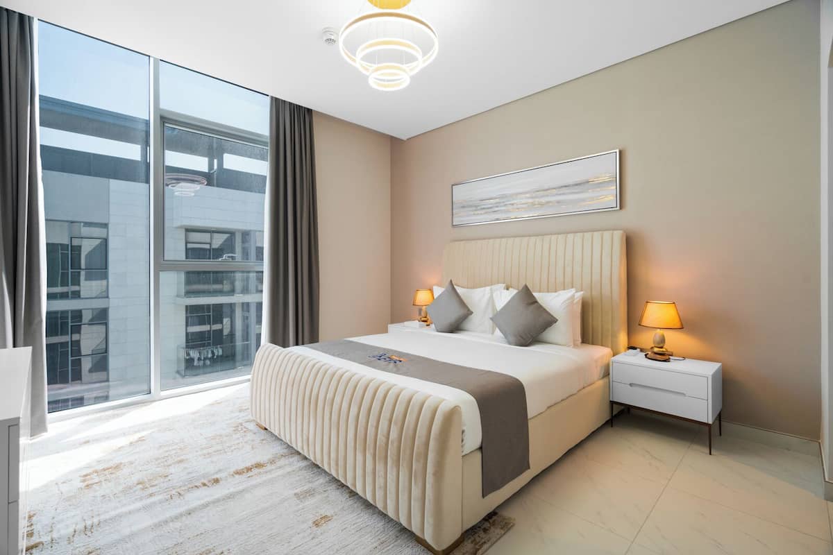 A modern hotel room with a large bed, a bedside table with lamps, a view of a building through large windows, and a ceiling light fixture.