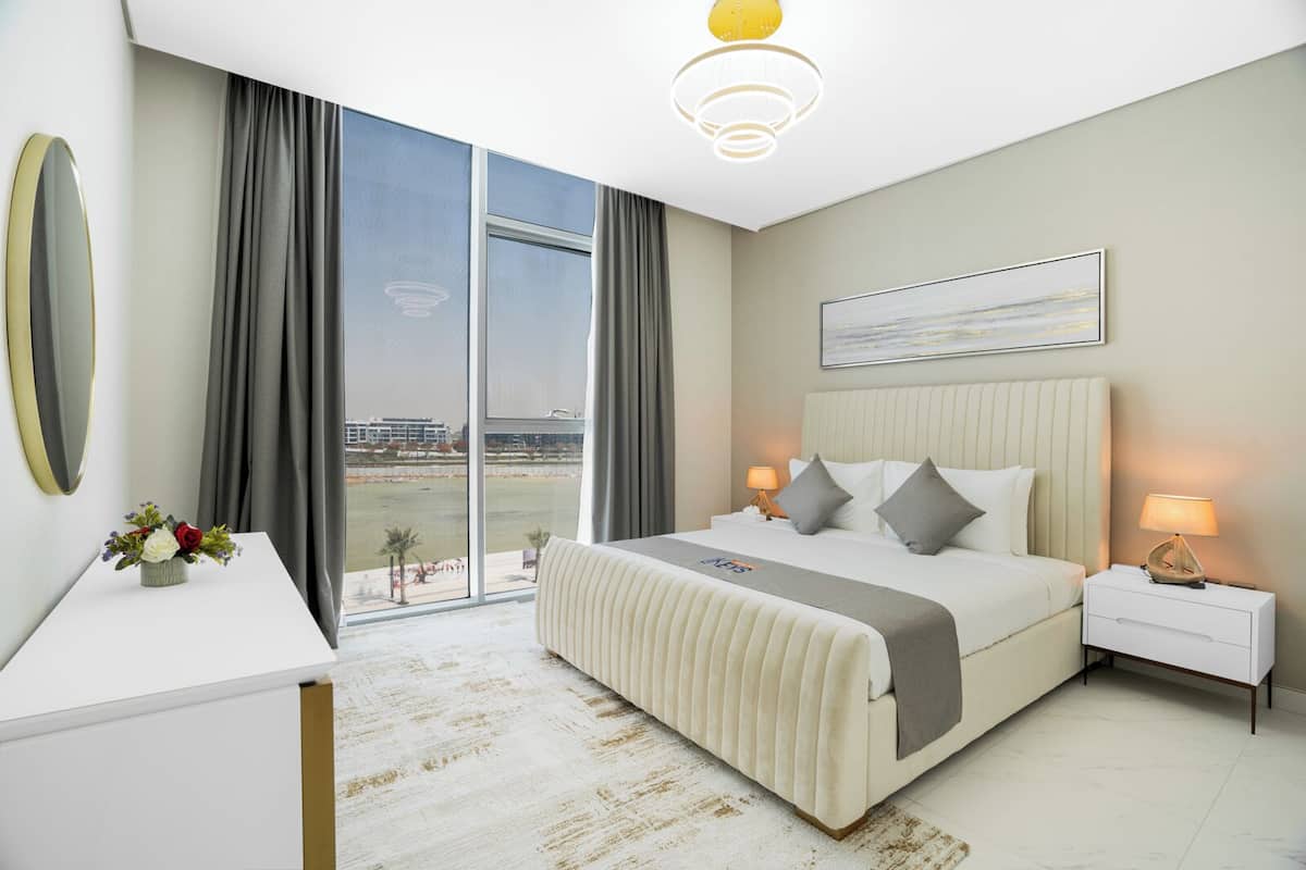 A modern hotel room with a large bed, a bedside table, a nightstand, a desk, a chair, and a view of the city.