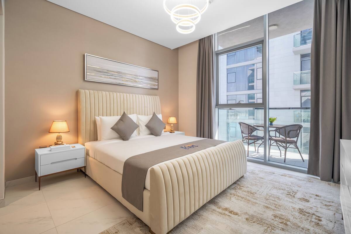 A modern hotel room with a large bed, bedside lamps, a view of the city through large windows, and a balcony with outdoor seating.