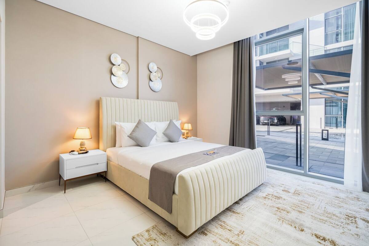 A modern hotel room with a large bed, bedside lamps, a view of the outside, and a unique wall art piece.