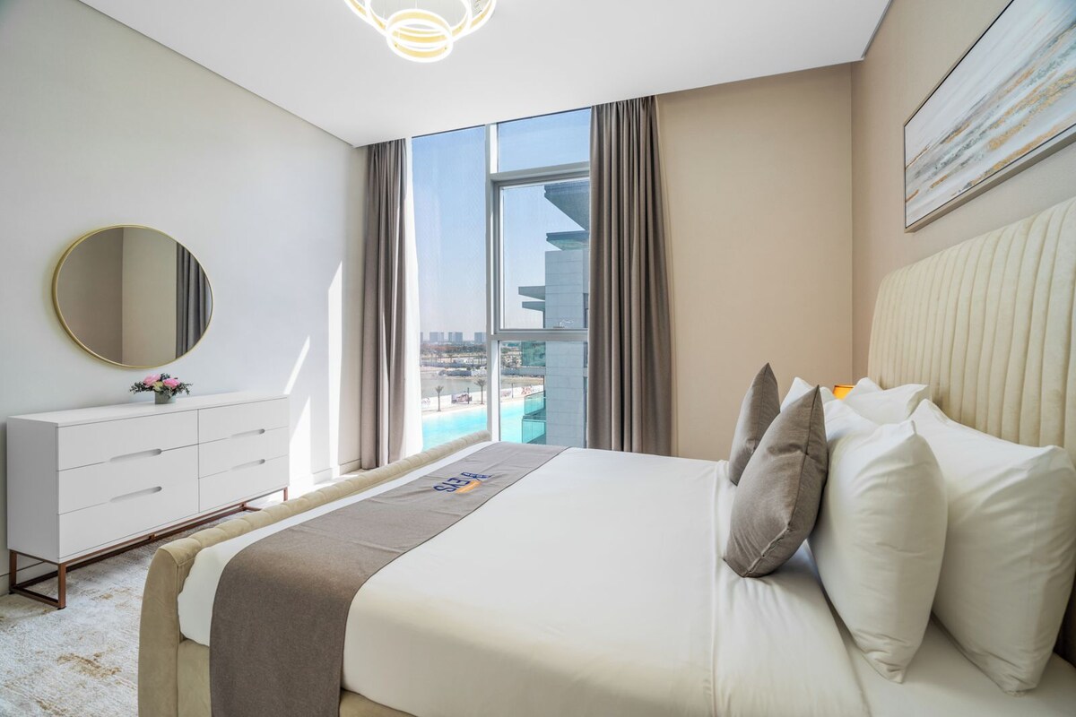 A modern hotel room with a large bed, a dresser, a round mirror, and a view of the cityscape.