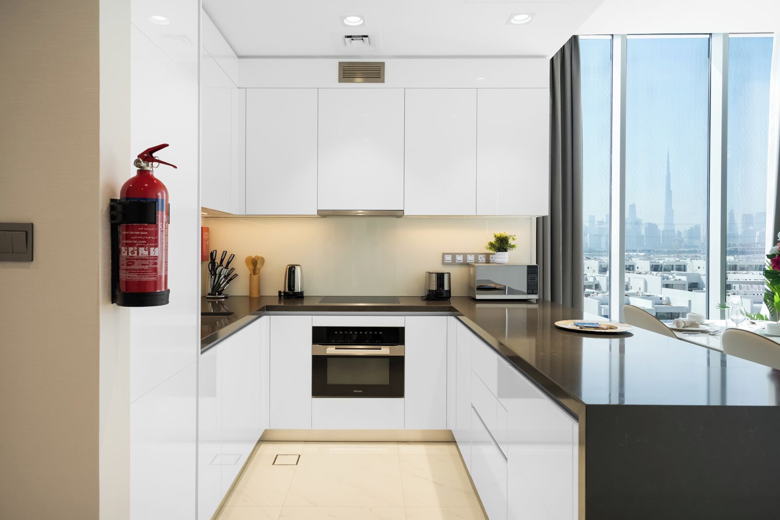 A modern kitchen with white cabinets, a built-in oven, a microwave, and a coffee maker. A fire extinguisher is mounted on the wall.