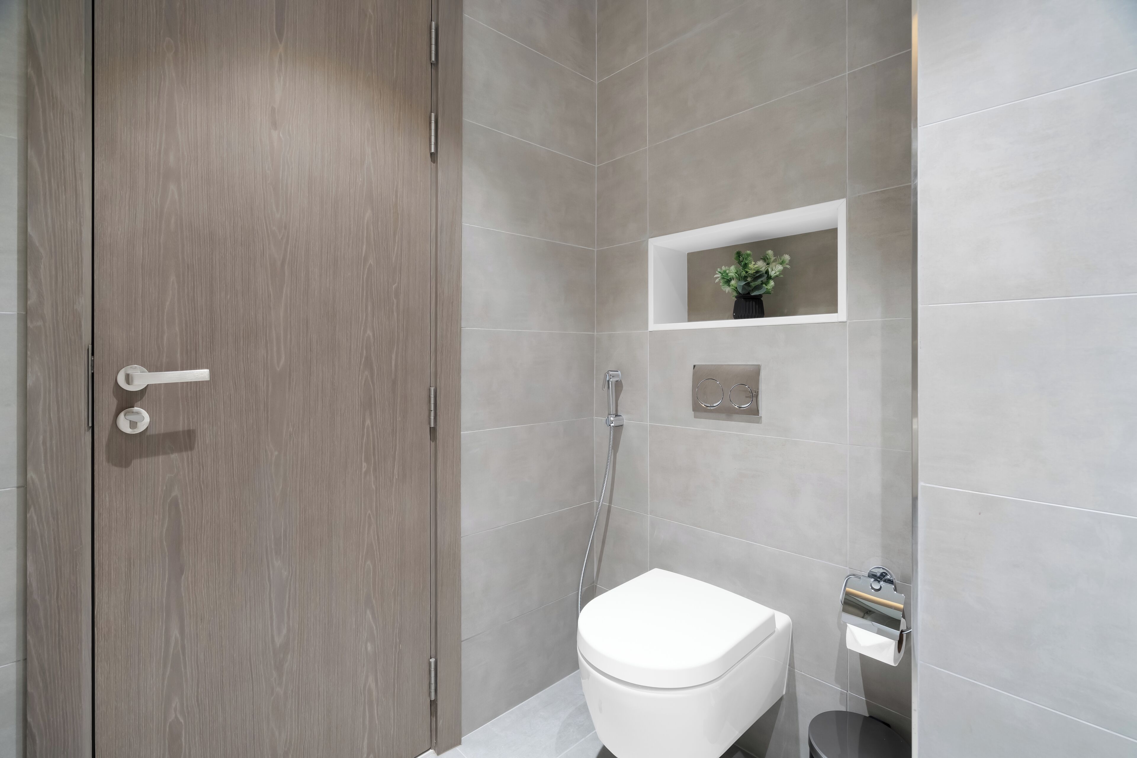 A modern bathroom with a wooden door, a white toilet, a shower area, and a small shelf with a plant.