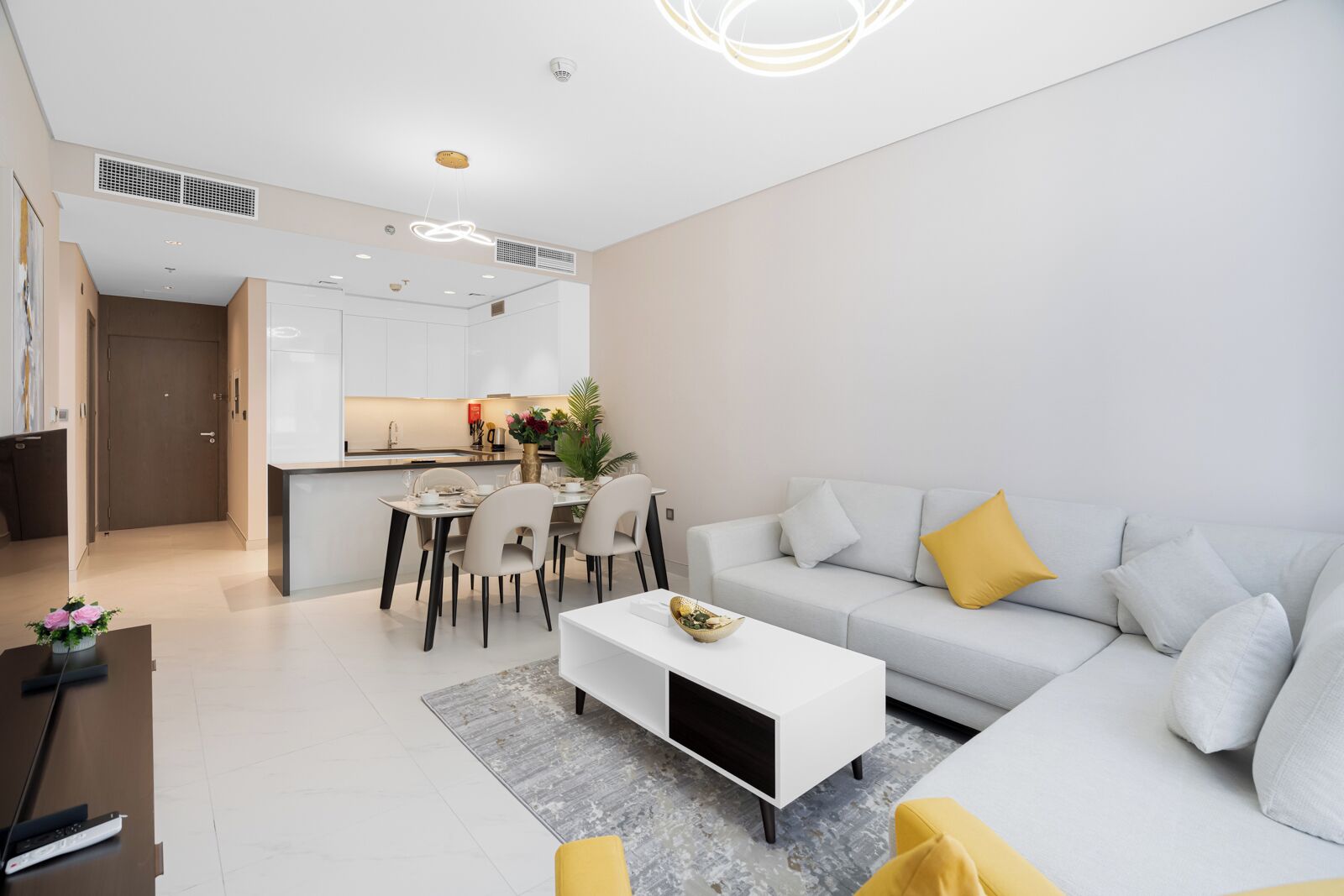 A modern living room with a grey sofa, yellow cushions, a white coffee table, and a dining area with a white table and chairs.