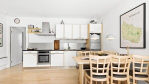 A modern kitchen with white cabinets, a built-in oven, and a dining area with wooden chairs and a table. - Gudhjem Søpark Lejl. 57 (Gudhjem)
