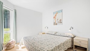 A bedroom with a large bed, two bedside tables, a wall-mounted lamp, and a window with curtains. - Gudhjem Søpark Lejl. 57 (Gudhjem)