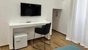 A compact room with a white desk, a black chair, a small white fridge, and a mounted TV. - Le Stanze Ritrovate (Trani)