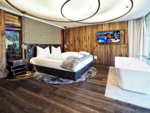 A modern bedroom with a large bed, a TV mounted on the wall, and a separate bathroom with a bathtub. - Wellness & Naturresort Reischlhof (Wegscheid)
