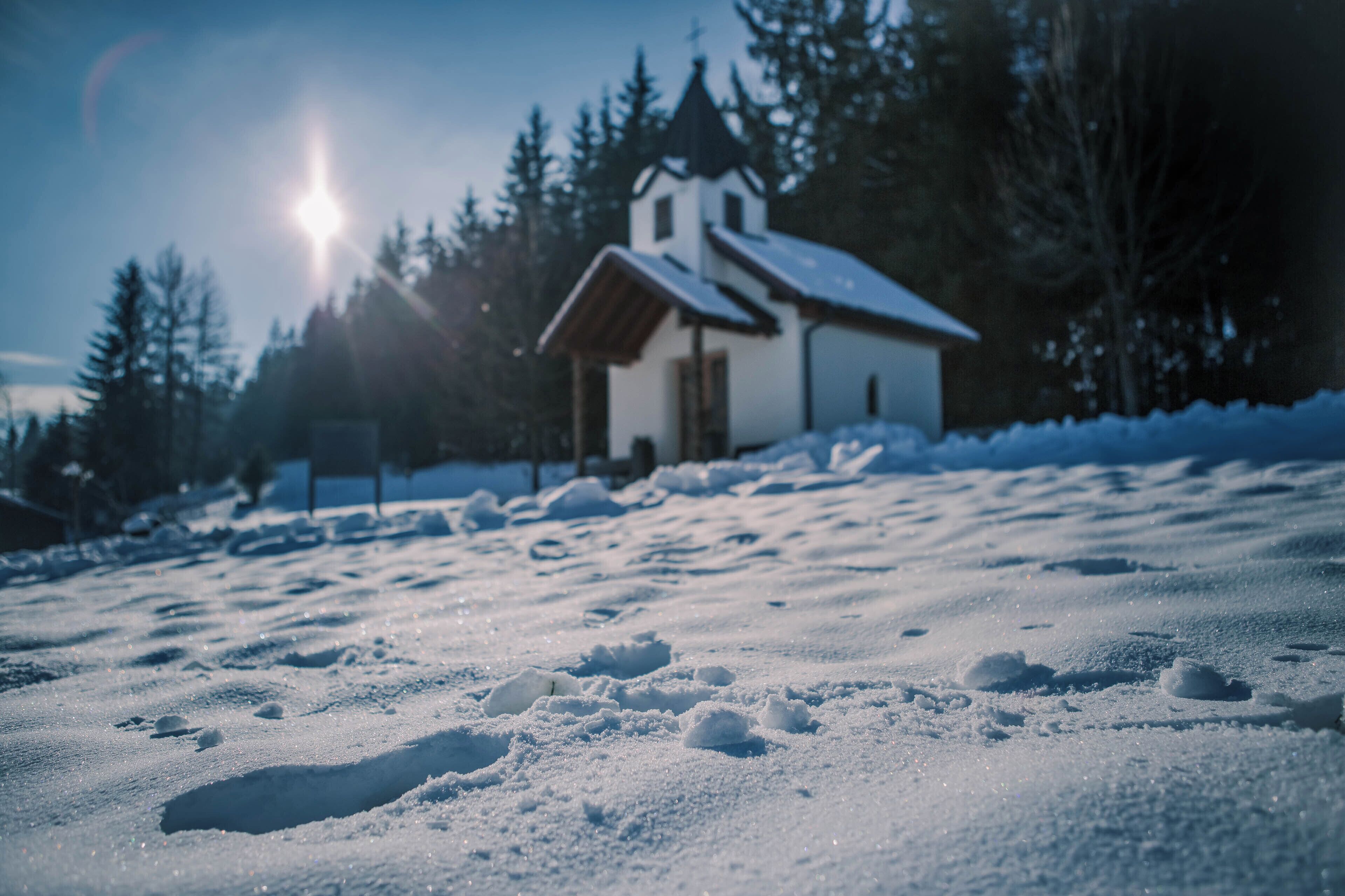 A snow-covered landscape with a small white building, likely a church, surrounded by trees.