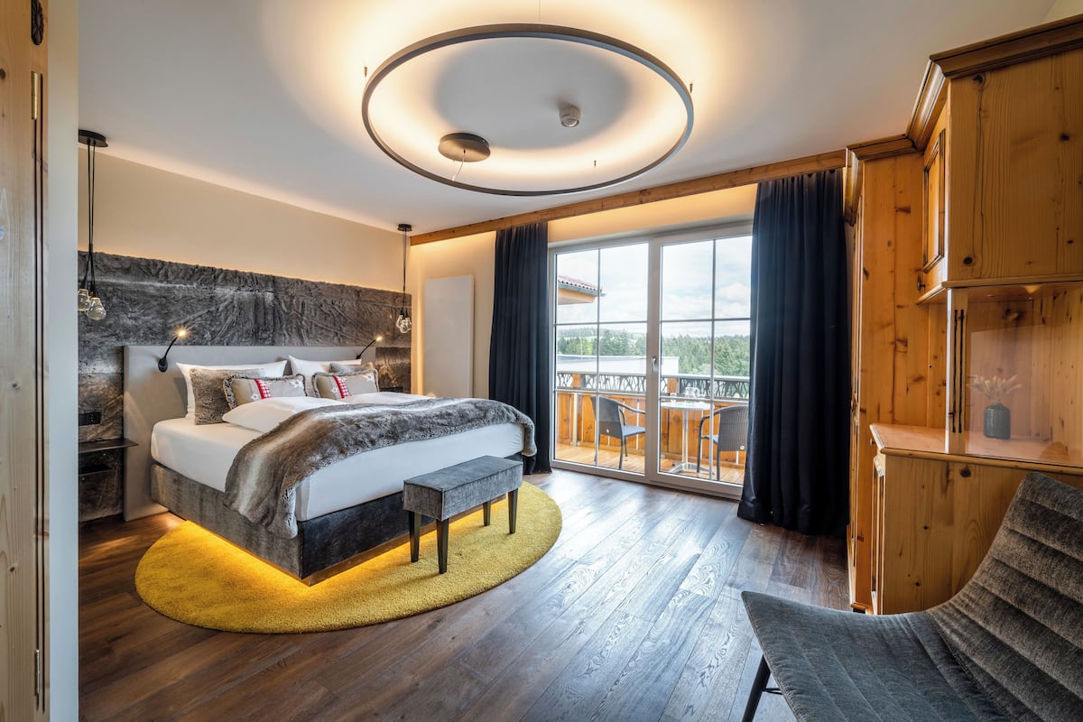 Une chambre moderne avec un grand lit, une tête de lit en bois, un luminaire circulaire au plafond et un balcon avec vue.