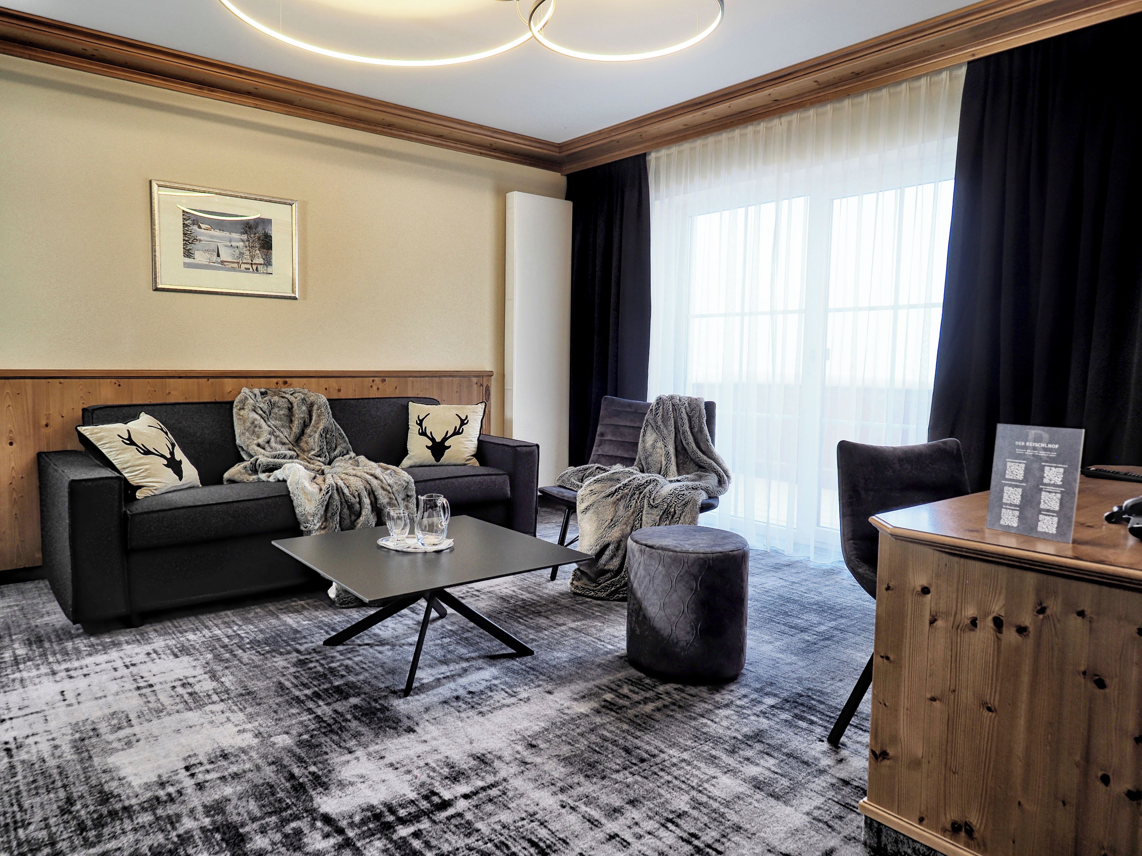 Une chambre d’hôtel moderne, dotée d’un canapé, d’un fauteuil et d’un pouf, d’une petite table avec des verres et d’une armoire en bois.