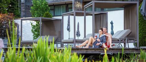 A couple relaxing on a deck with lounge chairs, overlooking a body of water and a building with a modern design.