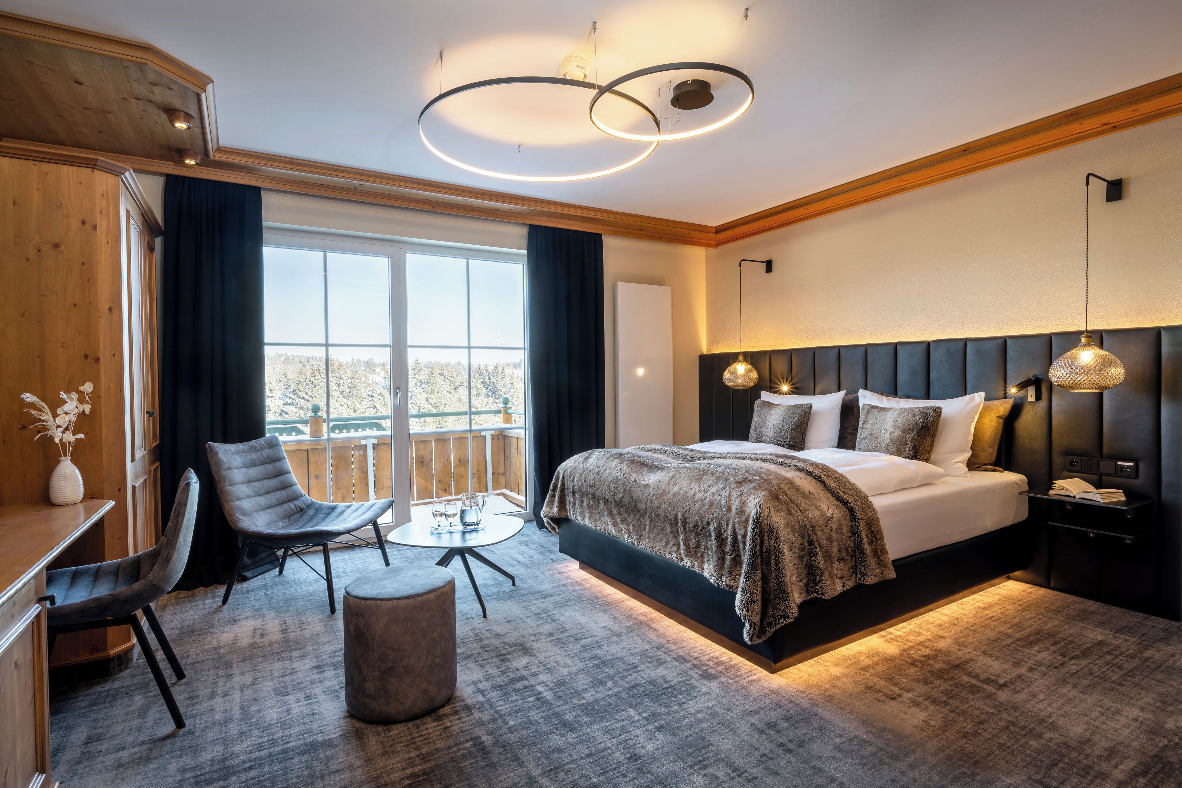 A modern hotel room with a large bed, a desk, two chairs, a small table, and a view of the outdoors.