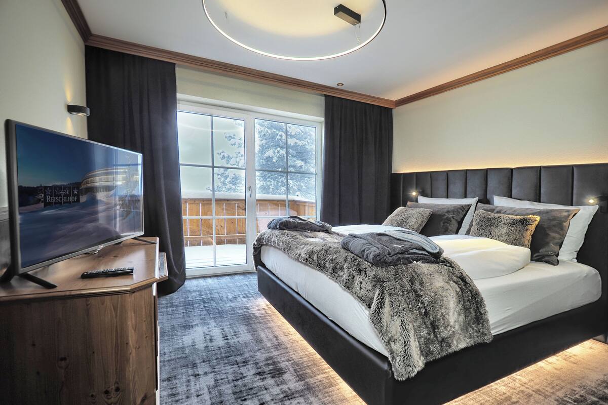 Une chambre moderne avec un grand lit, un téléviseur posé sur un meuble en bois et une vue sur un paysage enneigé par une grande fenêtre.