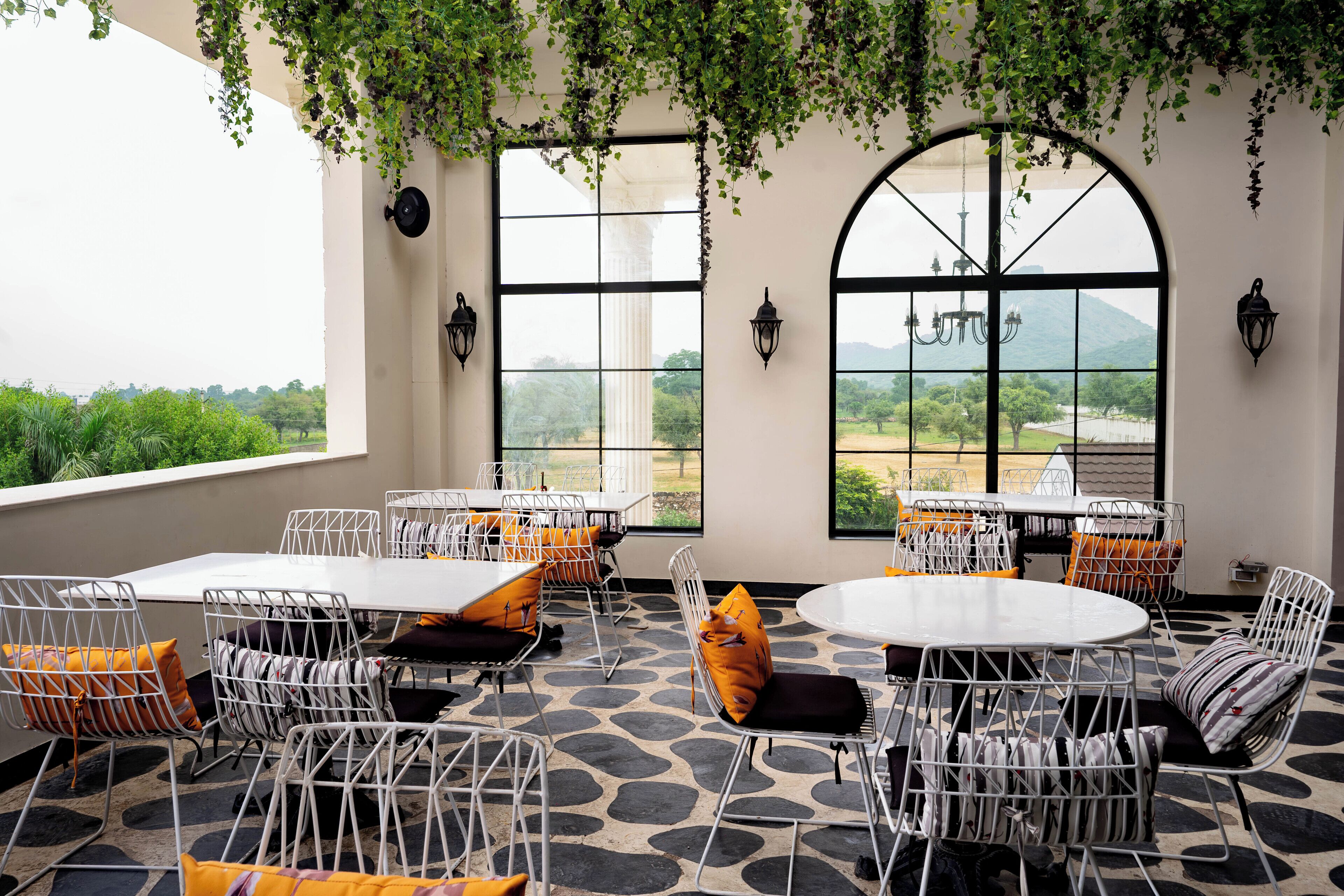 An outdoor dining area with white metal chairs and tables, large windows with views of greenery and mountains, and hanging plants above.