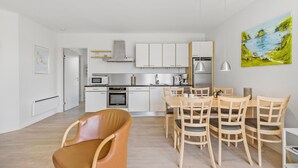 A modern kitchen with white cabinets, a microwave, oven, and a coffee maker. There is a dining area with wooden chairs and a table. - Gudhjem Søpark Lejl. 46 (Gudhjem)