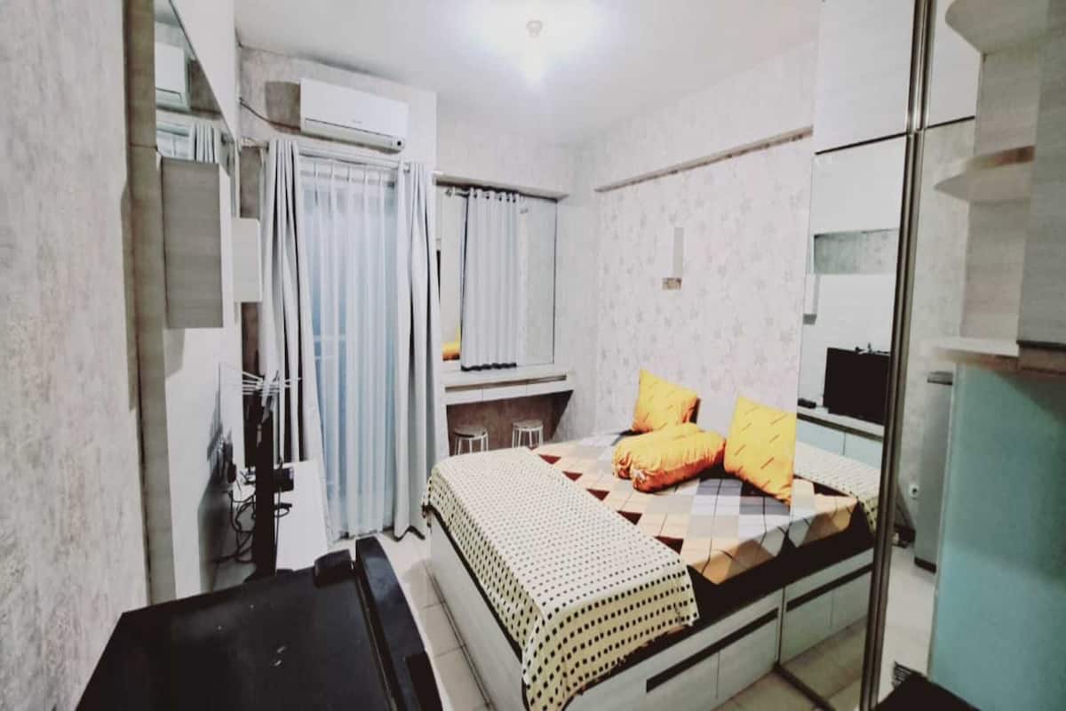 A small, compact bedroom with a bed, a desk, and a chair. There is a window with curtains, a radiator, and a small table.