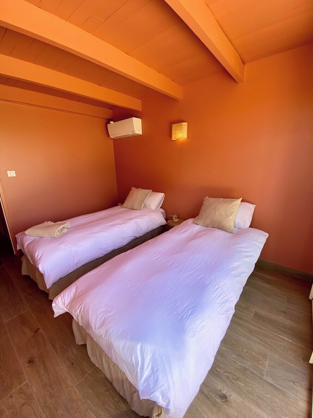 A room with two beds, wooden flooring, a ceiling fan, and a wall-mounted air conditioner.