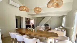 A spacious dining area with a large wooden table surrounded by six chairs, a kitchen in the background, and a staircase leading to an upper level.