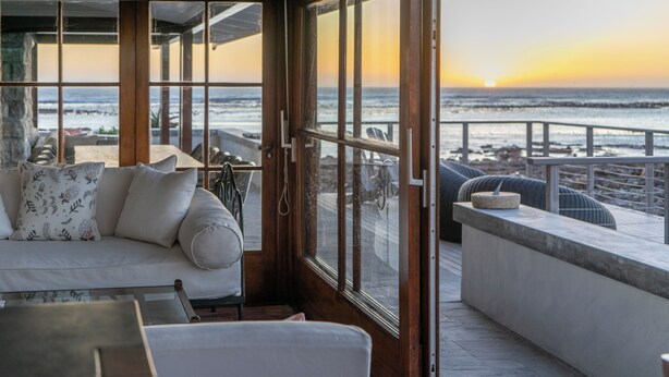 Una habitación costera con un amplio ventanal con vistas al océano al atardecer, un sofá con cojines estampados y una pequeña mesa.