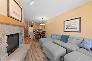 A living room with a stone fireplace, a gray sectional sofa, and a dining area with wooden cabinets and a refrigerator. - Ideal Location in Keystone Base Area, Private Balcony w/ Pool & Mountain View (Keystone)