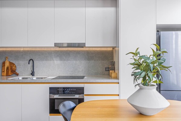 A modern kitchen with white cabinets, a stainless steel sink, an oven, and a wooden dining table with a white vase. - Executive Beachside Getaway – Cotton Tree (Sunshine Coast)